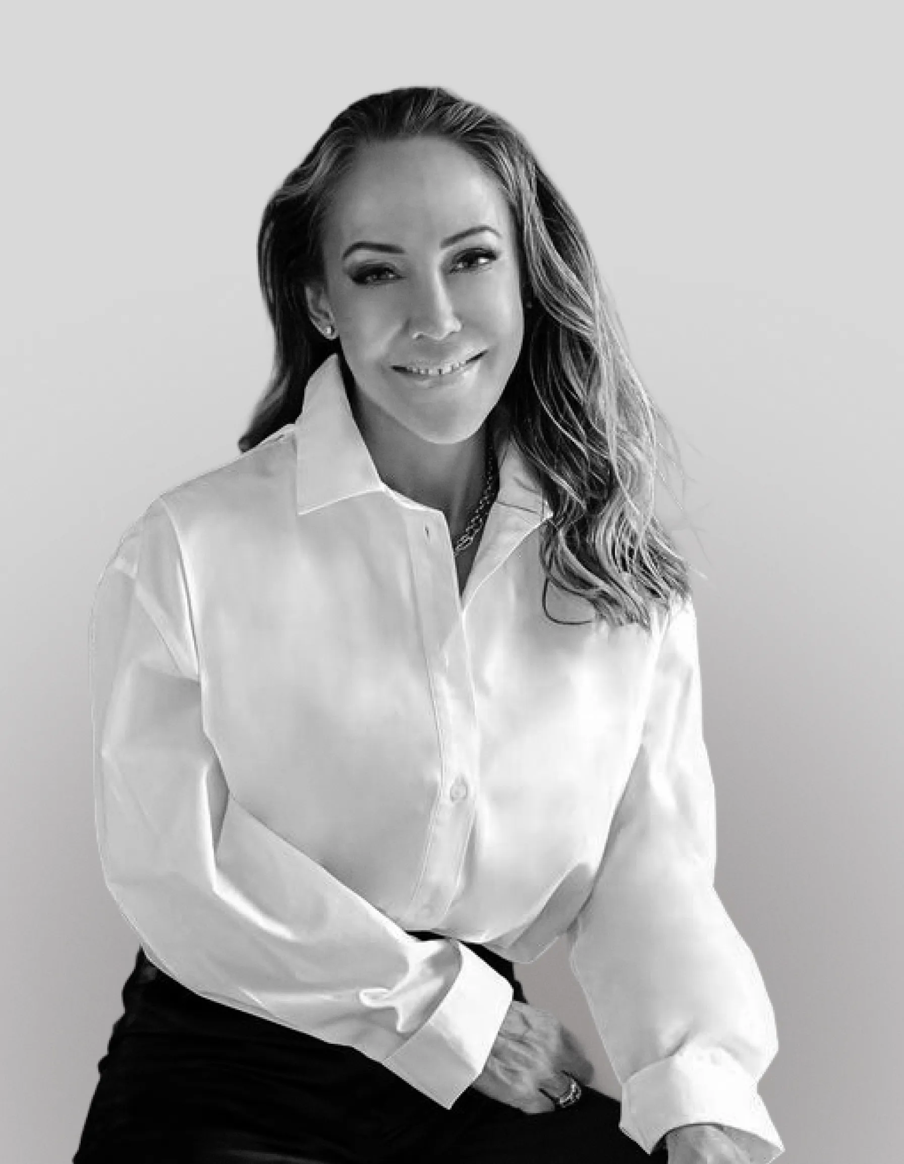Smiling woman with long hair wearing a white button-up shirt and dark pants, seated against a plain background.