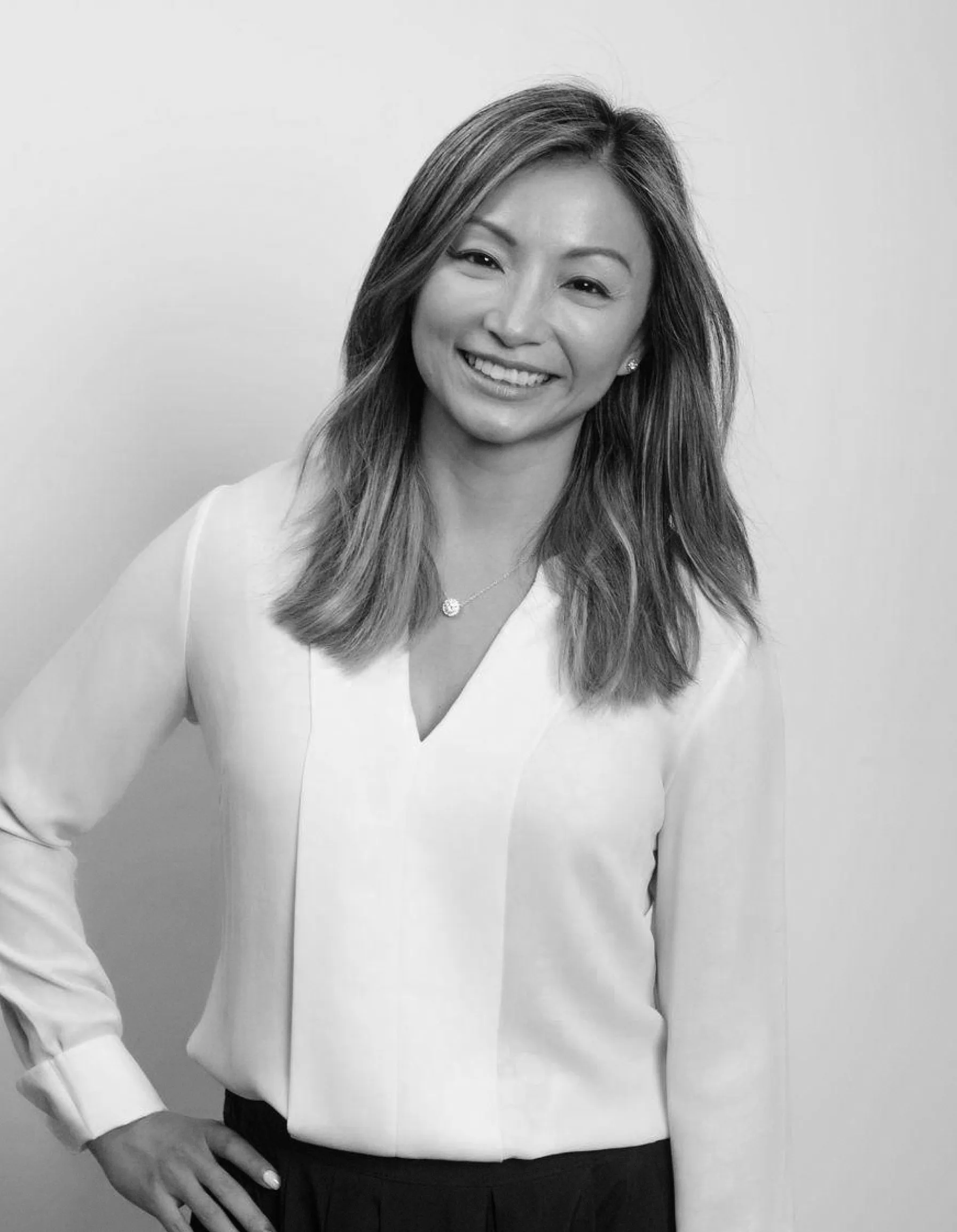 Smiling woman with long hair wearing a white blouse and black pants, standing with one hand on her hip.