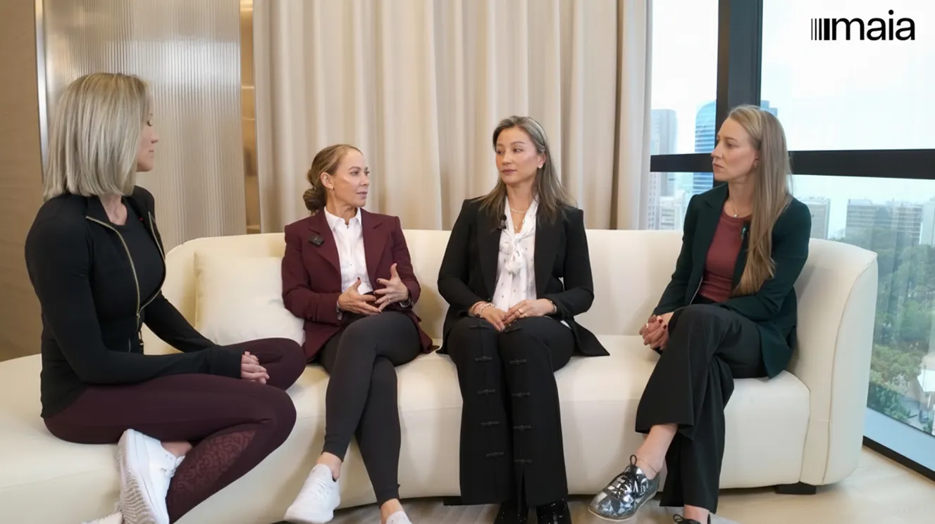 Four women seated on a white couch in a modern office with large windows, engaged in a discussion.