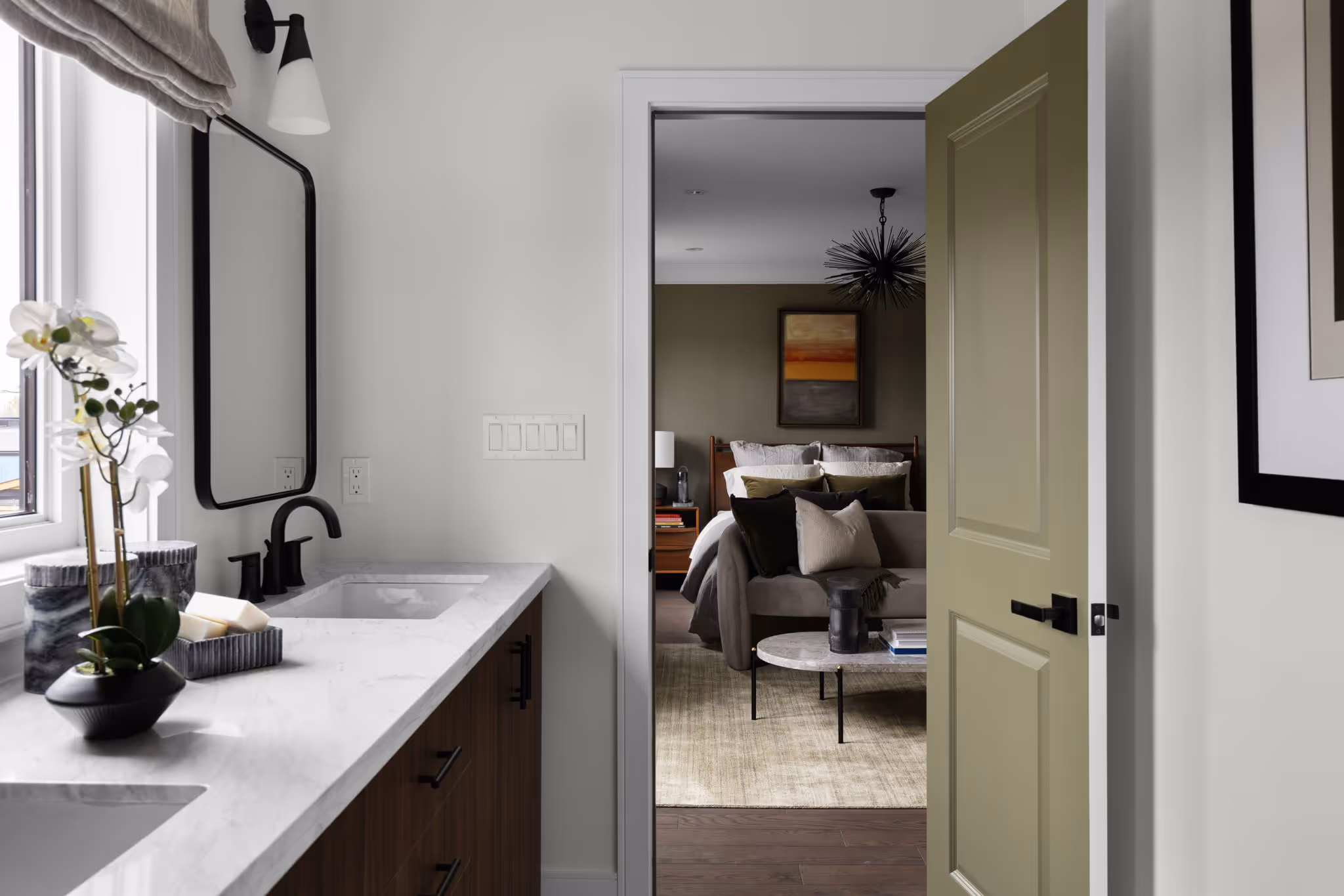Modern bathroom vanity with marble countertop and black fixtures next to an open door revealing a bedroom with a couch, coffee table, and bed.