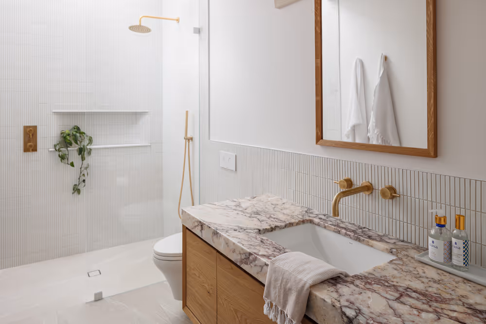 Modern bathroom with marble countertop, gold faucet, wooden framed mirror, and walk-in shower with gold showerhead and trailing plant.