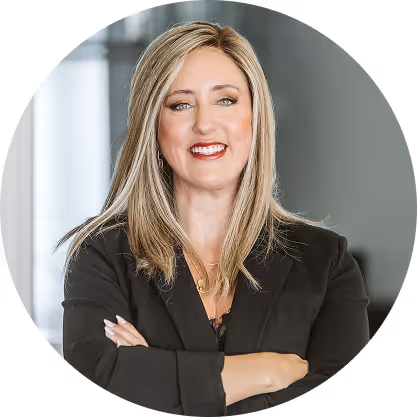 Confident woman with blonde hair wearing a black blazer, smiling with arms crossed against a blurred gray background.