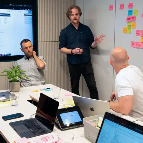 383 Group team members working in a meeting room in front of a digital screen and with post-it notes stuck to a wall. 