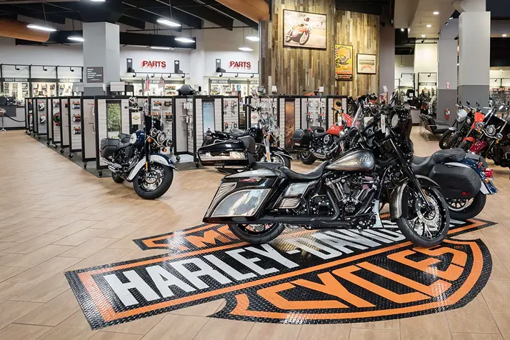 A Harley Davidson showroom with two bikes in the forefront and many more behind. The bikes in the fore are parked on a floor vinyl of the Harley logo. 