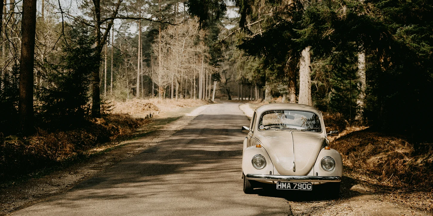 A classic Volkswagen Beetle in beige parked up at the side of a secluded road in a woodland area. 