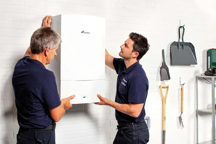 Two technicians in navy shirts installing a white Worcester Bosch boiler on a white brick wall.