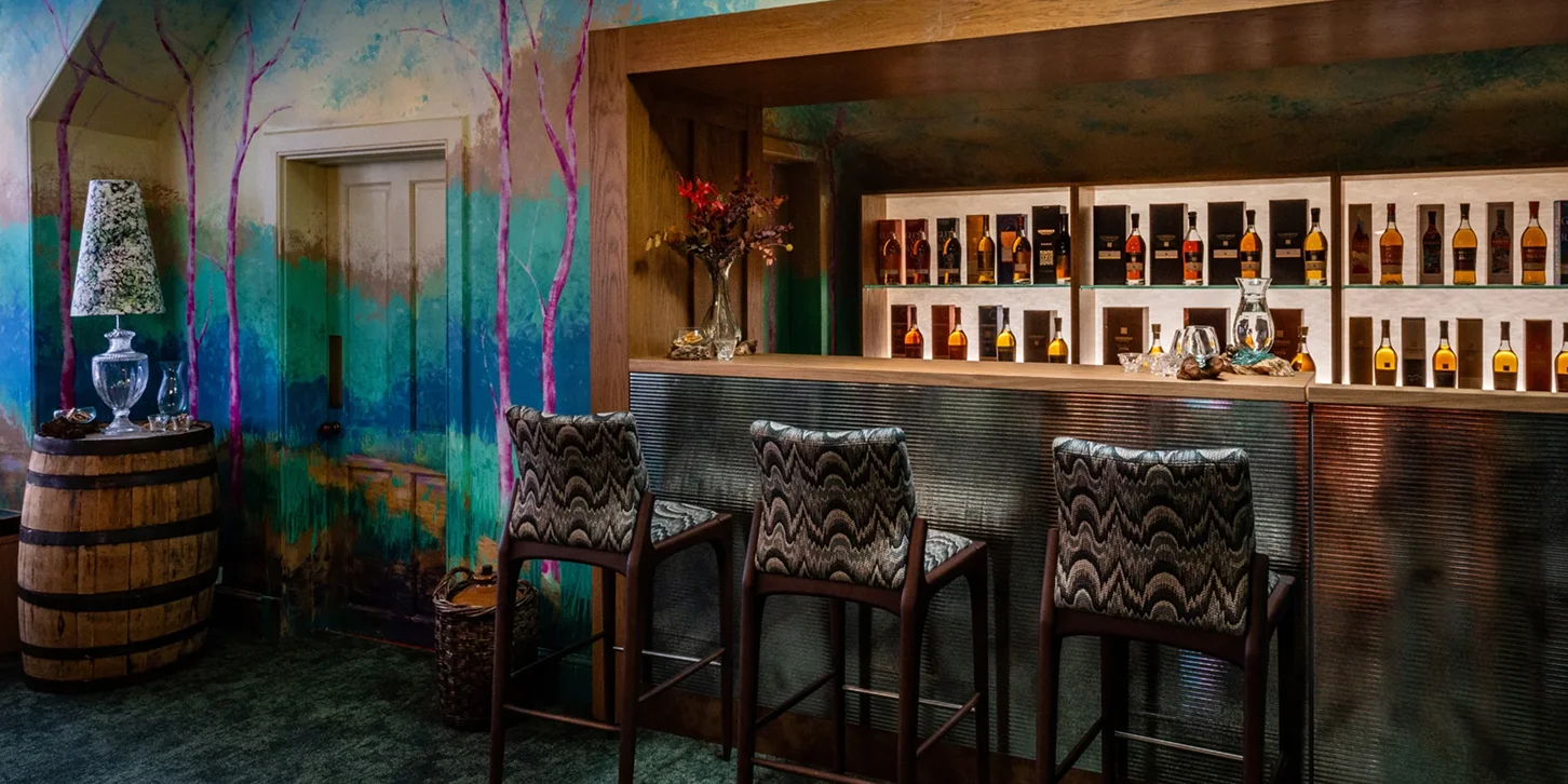 Cozy bar interior with three patterned high chairs, colorful tree mural walls, wooden barrel table with lamp, and shelves of liquor bottles.