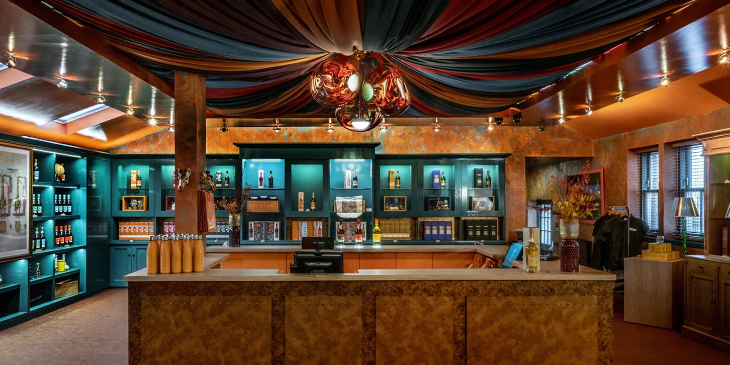 Interior of a warmly lit retail shop with teal shelves displaying bottles and orange counters beneath a draped fabric ceiling and decorative hanging lights.