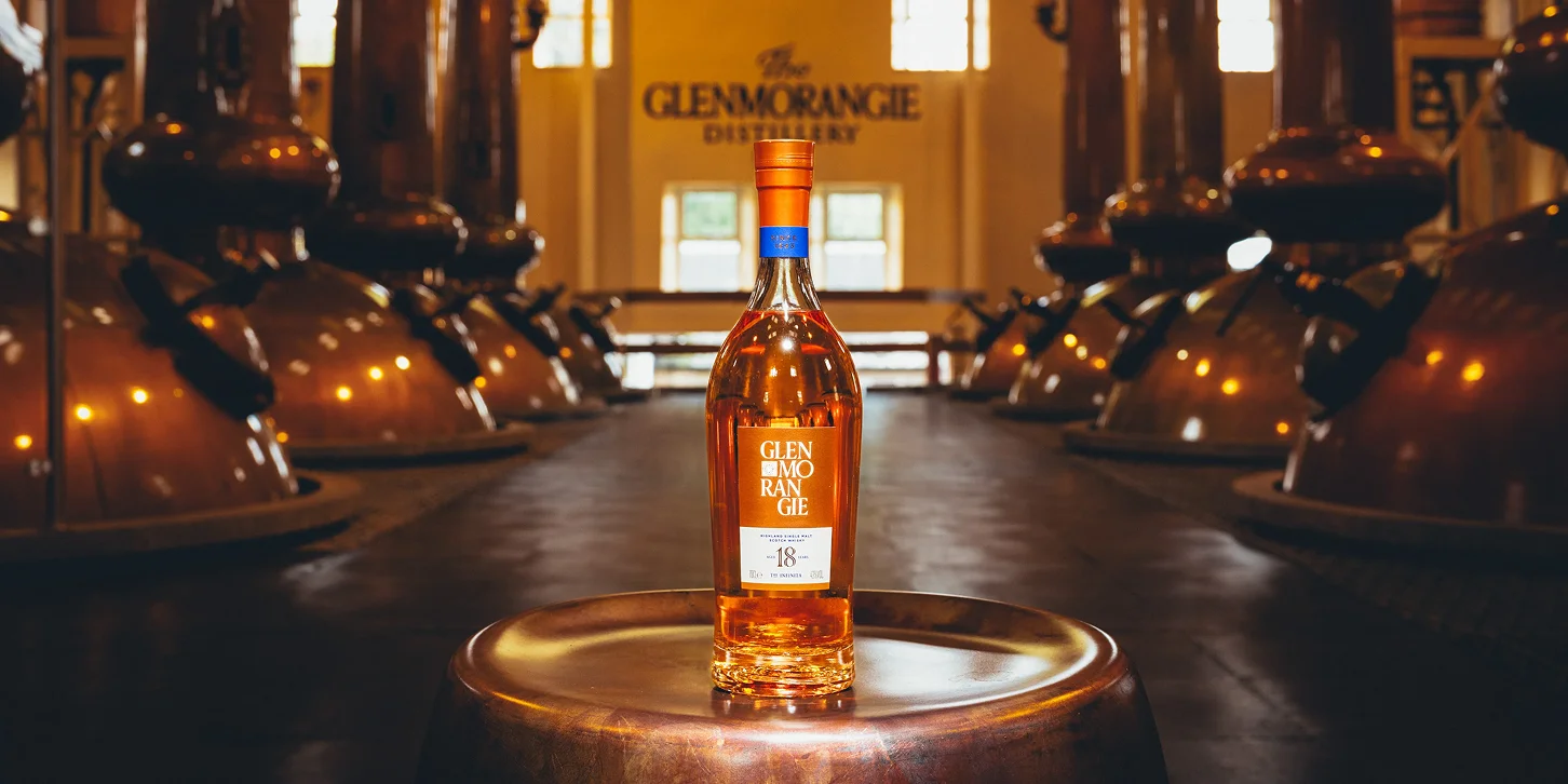 Bottle of Glenmorangie 18-year-old single malt Scotch whisky displayed on a copper table inside the Glenmorangie distillery with copper stills in the background.