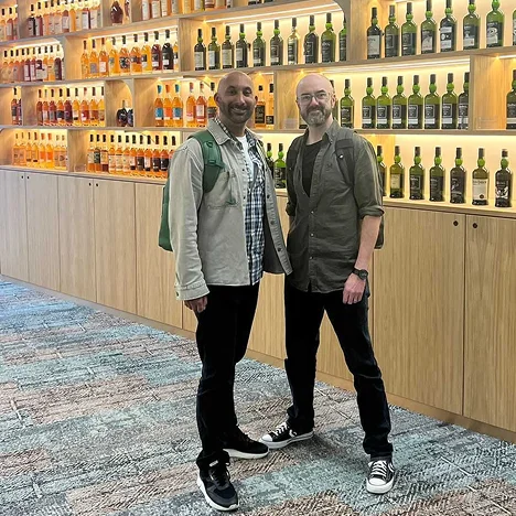 Two men standing and smiling in front of well-lit wooden shelves filled with bottles of whiskey.