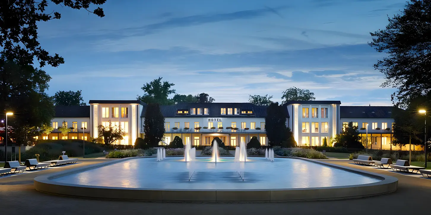 Lit hotel building at dusk seen from across a large fountain with water jets and surrounded by benches and trees.