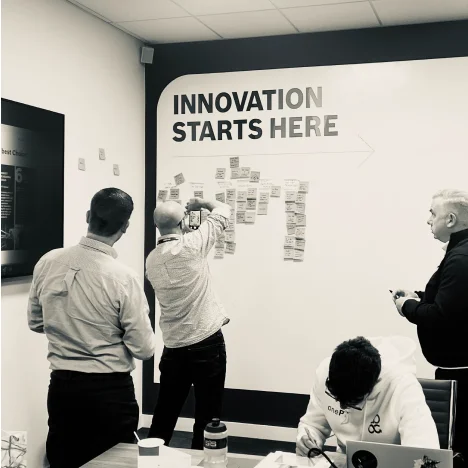 Four people collaborating in a meeting room with a wall that reads 'INNOVATION STARTS HERE' and sticky notes arranged on it.