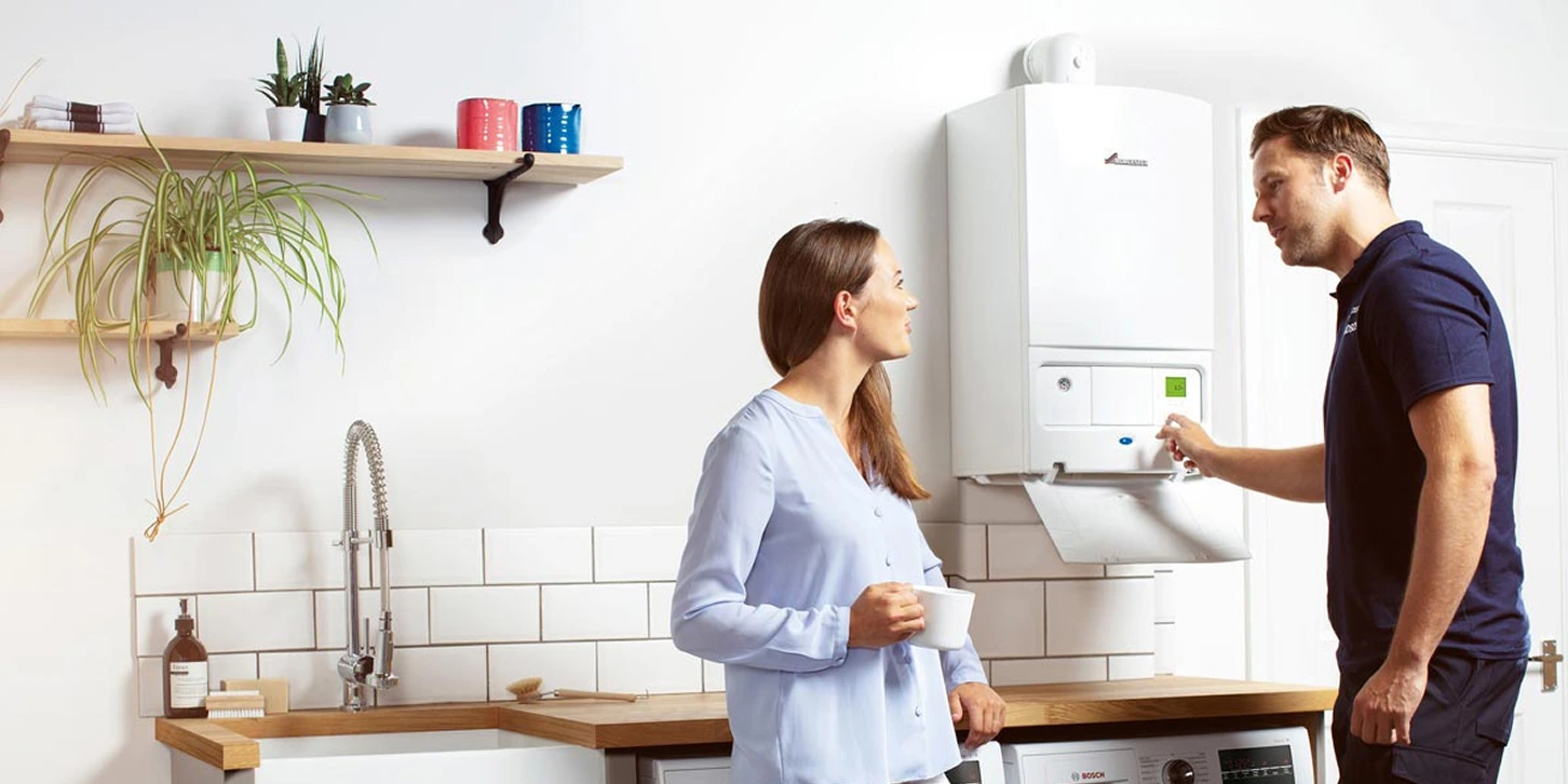 Technician adjusting settings on a wall-mounted boiler while a woman in a light blue blouse watches holding a white mug in a modern kitchen.