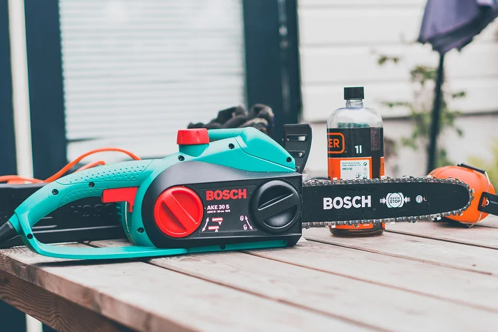 Bosch AKE 30 S electric chainsaw placed on a wooden table with a bottle and orange protective gear in the background.