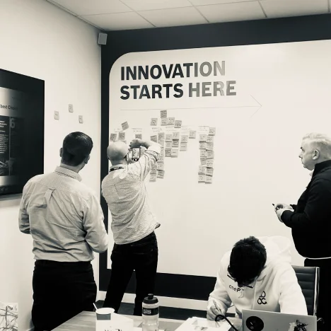 Four people collaborate in a meeting room with a wall reading 'INNOVATION STARTS HERE' and sticky notes arranged on the wall.