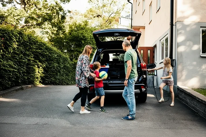 Family of four packing a black SUV with the trunk open in a driveway near a house surrounded by greenery.