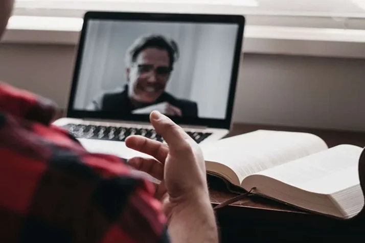 Person gesturing with hand during a video call on a laptop with an open book nearby.
