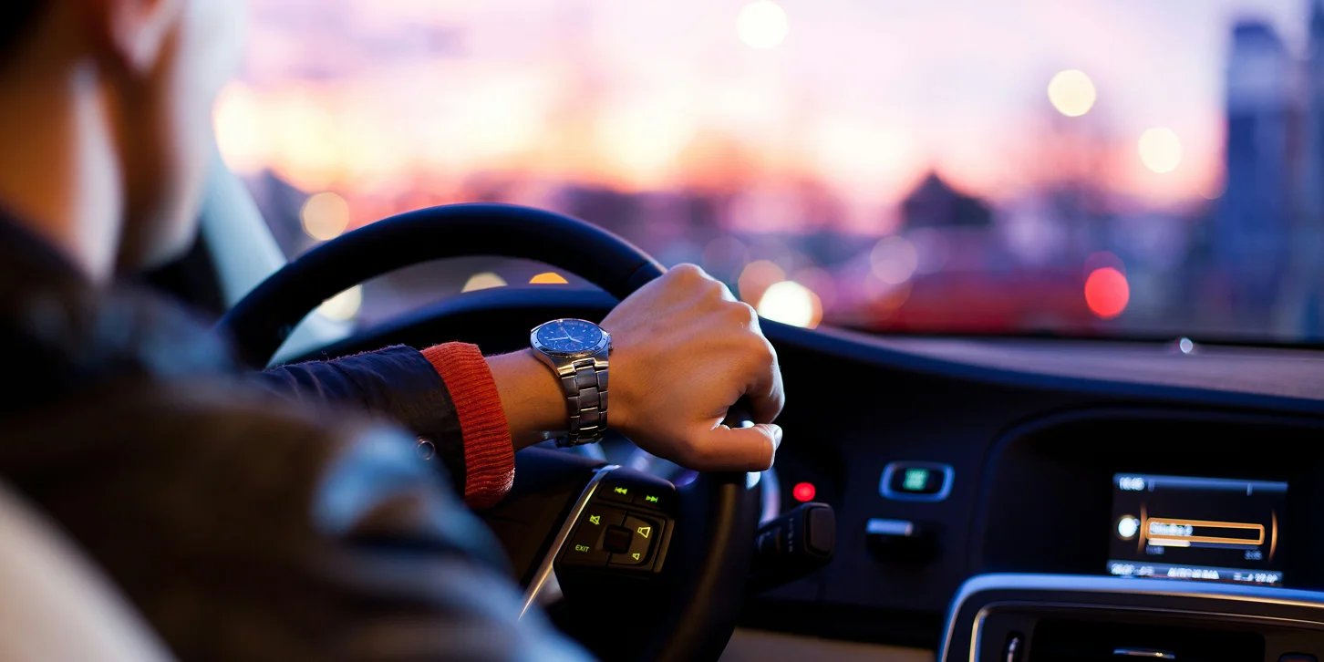 Person driving a car at sunset with a watch on their wrist and dashboard lights visible.
