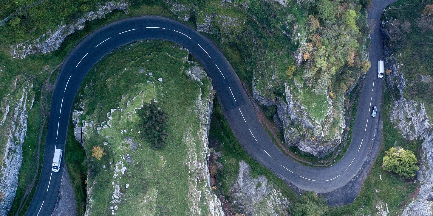 Aerial view of a winding mountain road surrounded by green rocky terrain with three vehicles driving along it.