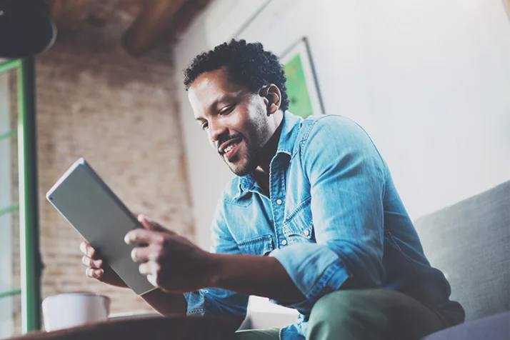 Man in blue denim shirt sitting on a couch and looking at a tablet with a smile.