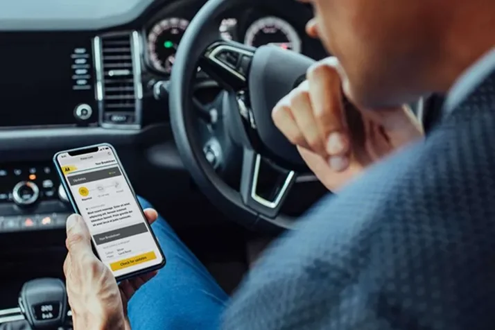 Person seated in a car, holding a smartphone displaying a vehicle inspection app, with the car dashboard and steering wheel in view.