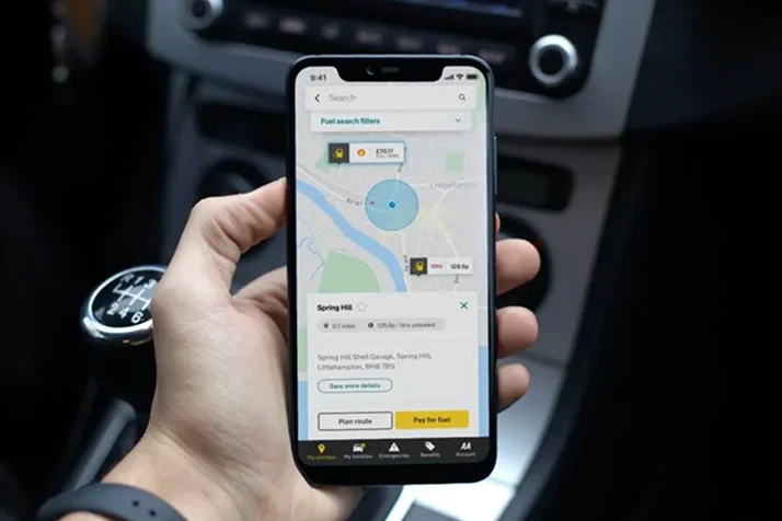 Hand holding smartphone displaying a map and fuel search results for Spring Hill Shell Garage inside a car with manual gear shift.