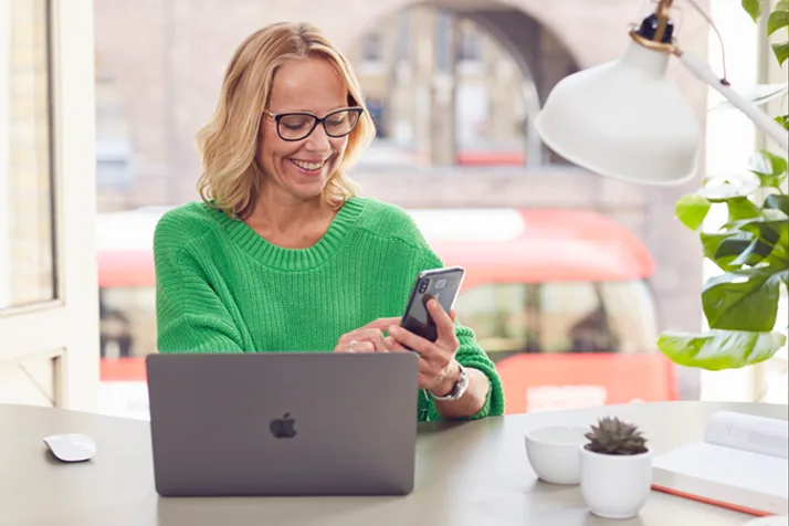 Smiling woman in green sweater using smartphone at a desk with a laptop and plants near a window.