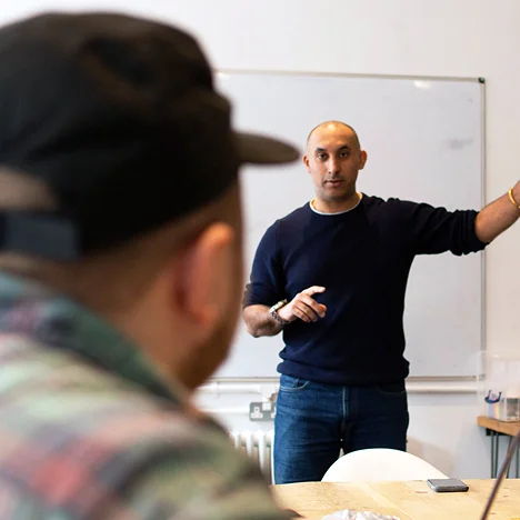 Man in dark sweater giving a presentation or lecture to a seated audience in a room with a whiteboard.