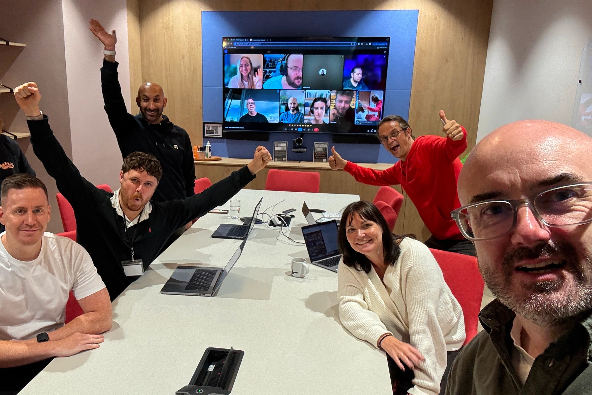 A group of six people enthusiastically posing around a conference table with laptops, while a virtual meeting with eight participants is displayed on a large screen behind them.