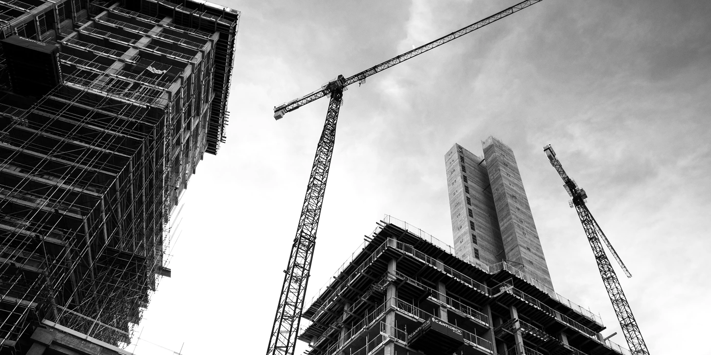 A black and white shot from a construction site, shot from the ground looking up a high rise frames of buildings being built and with cranes working on site. 