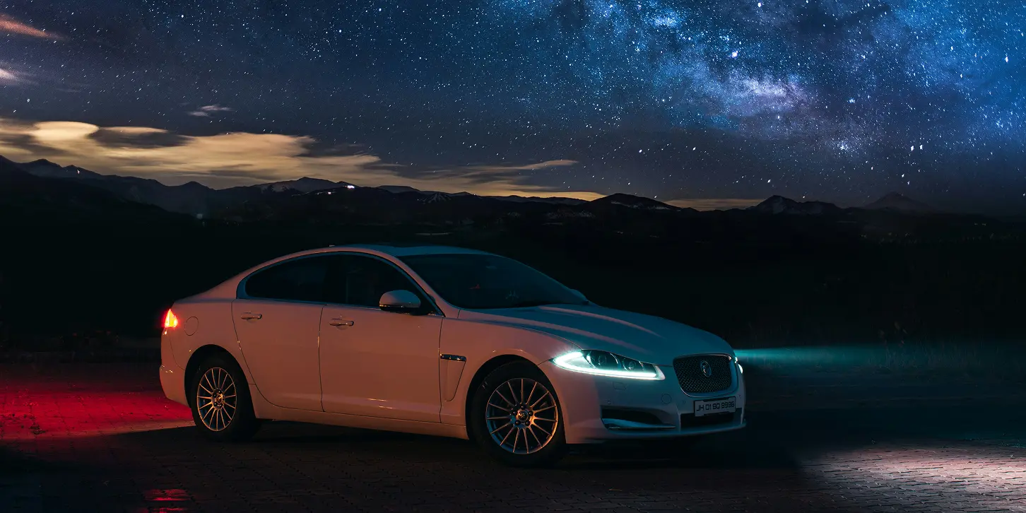White luxury sedan parked at night with headlights and rear lights on under a starry sky.