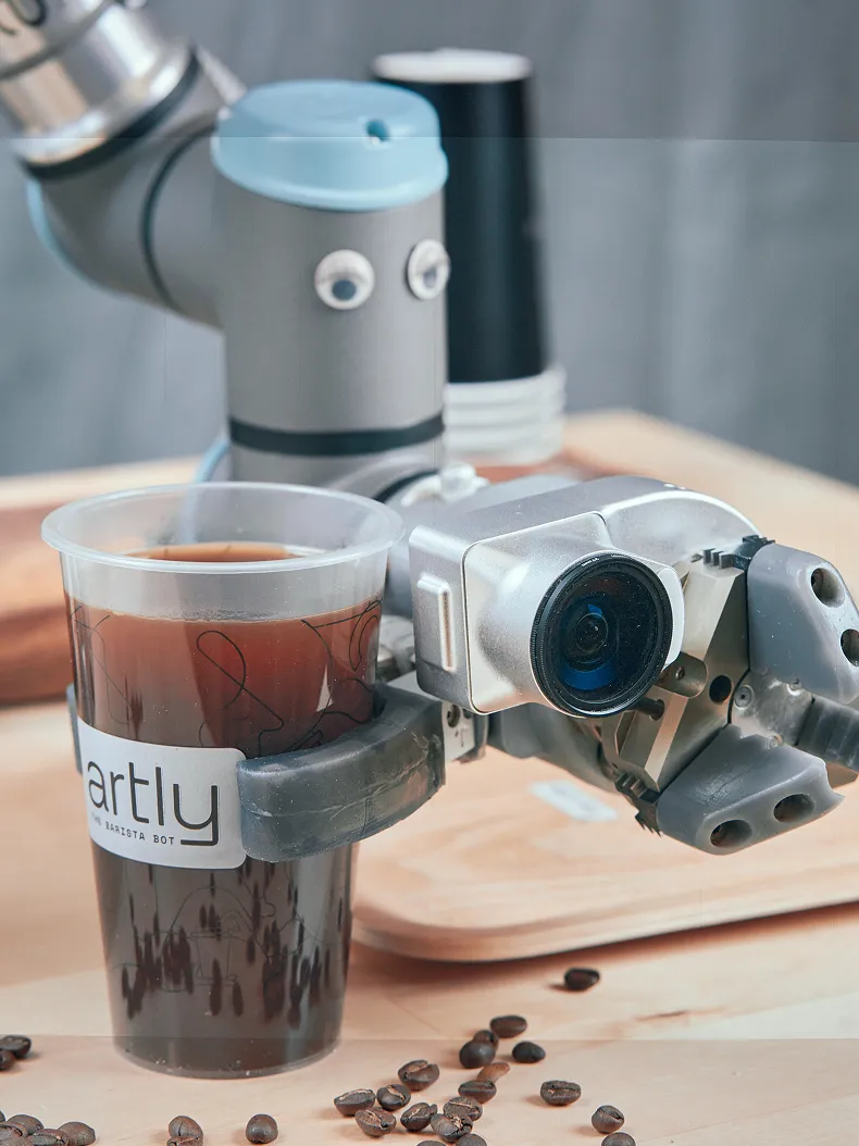 Artly robot arm with integrated vision sensor gripping a branded coffee cup, surrounded by scattered coffee beans.