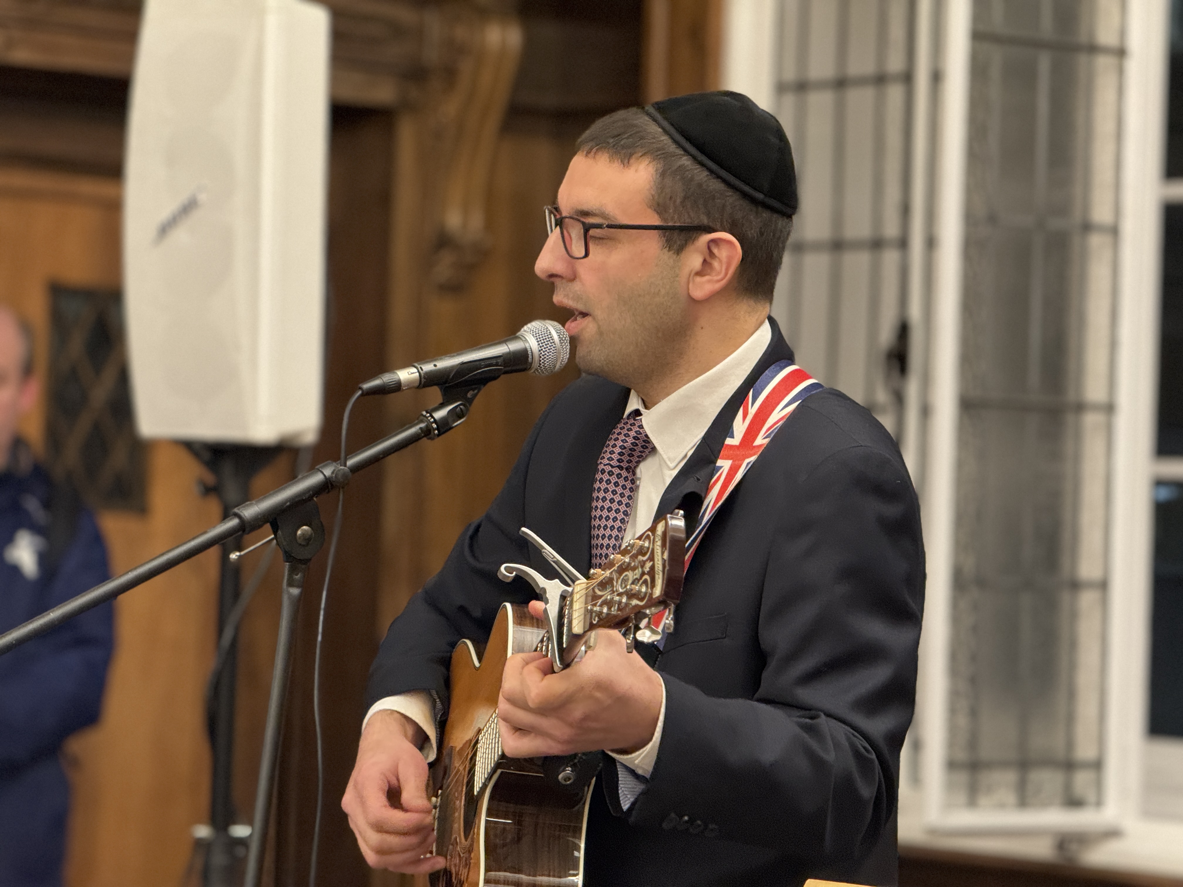 Musician in suit and kippah singing and playing acoustic guitar into microphone