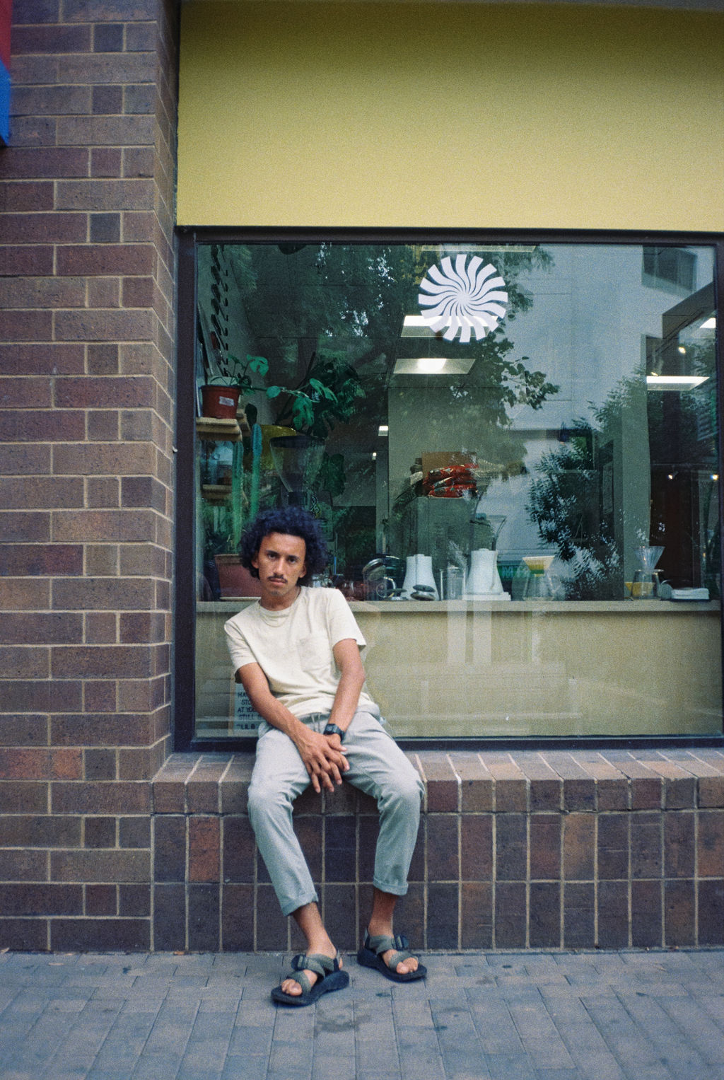 Man with curly hair wearing a beige t-shirt and gray pants sitting on a brick ledge outside a glass window.