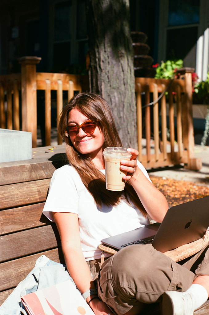Woman with sunglasses sitting on a wooden bench outdoors, holding an iced coffee and using a laptop.