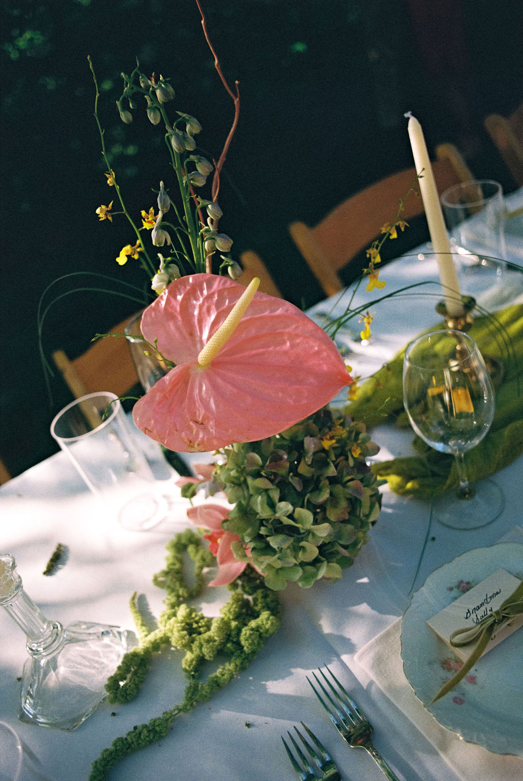 Table setting with floral centerpiece featuring pink anthurium, green hydrangeas, and yellow flowers, alongside candles, wine glasses, and place card.