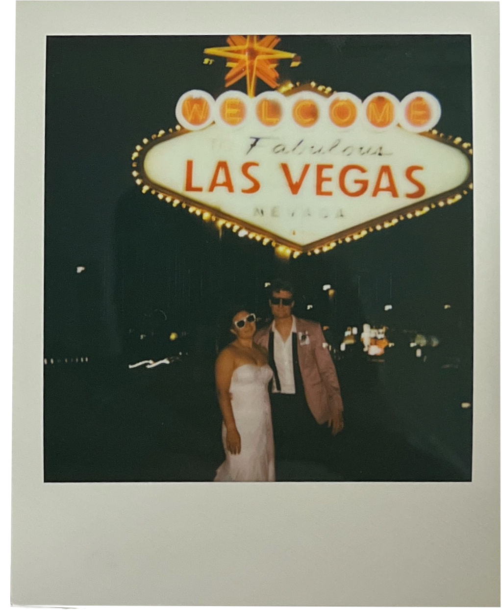 Couple wearing sunglasses posing in front of the illuminated Welcome to Fabulous Las Vegas sign at night.