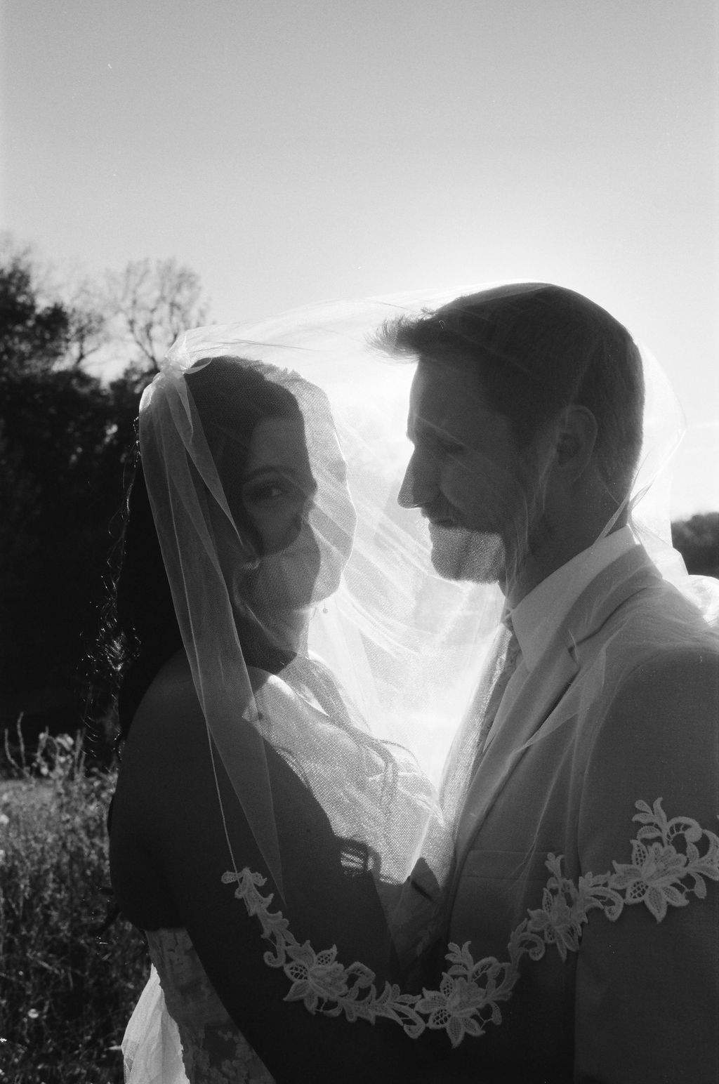 Black and white photo of a bride and groom smiling at each other under the bride’s veil outdoors at sunset.