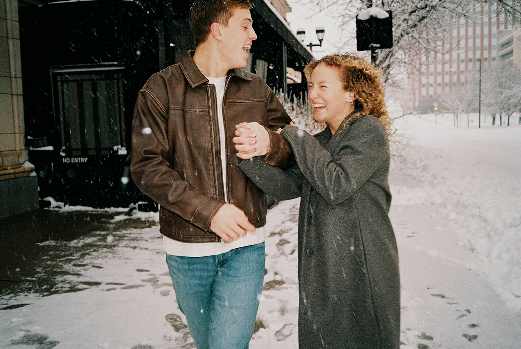 Young man and woman laughing and holding hands while walking on a snowy sidewalk in a city.
