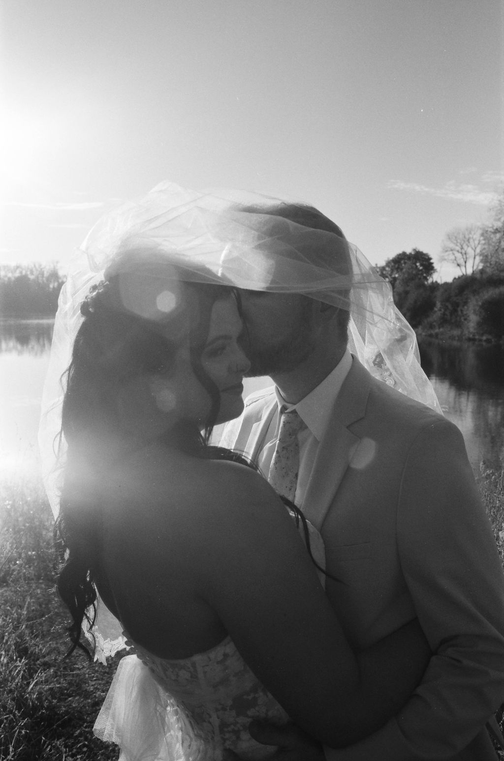 Black and white photo of a bride and groom embracing outdoors with a lake and trees in the background, the groom kissing the bride's temple under her veil.