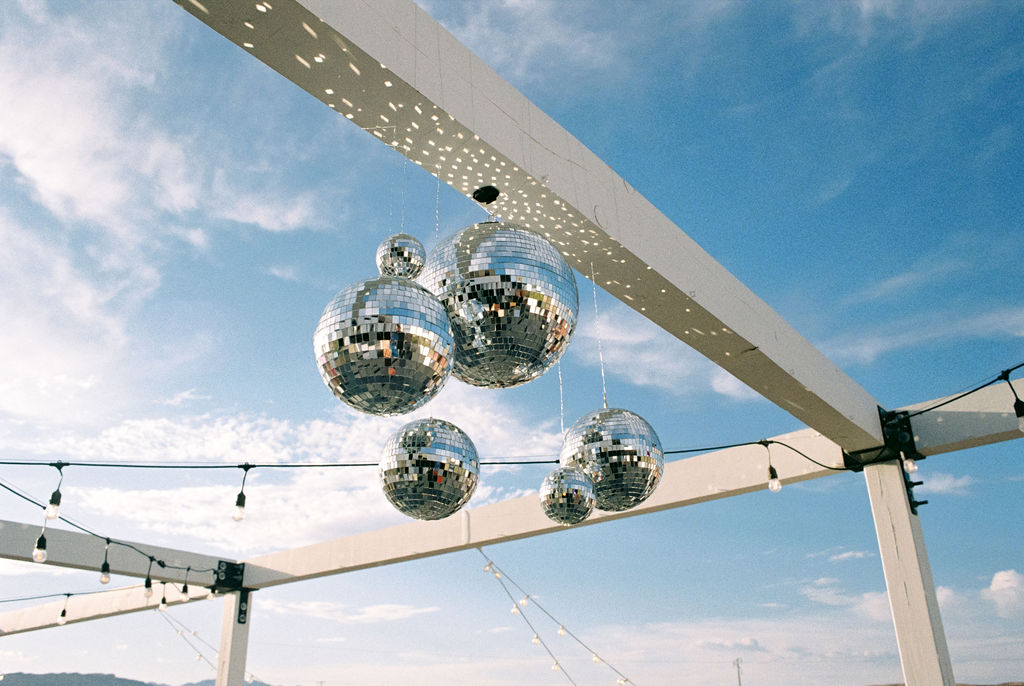 Cluster of five mirrored disco balls hanging from a white pergola against a blue sky with string lights.