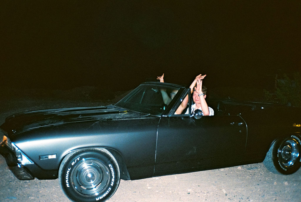 Two people in a black convertible car at night, with one person raising their hands outside the car.