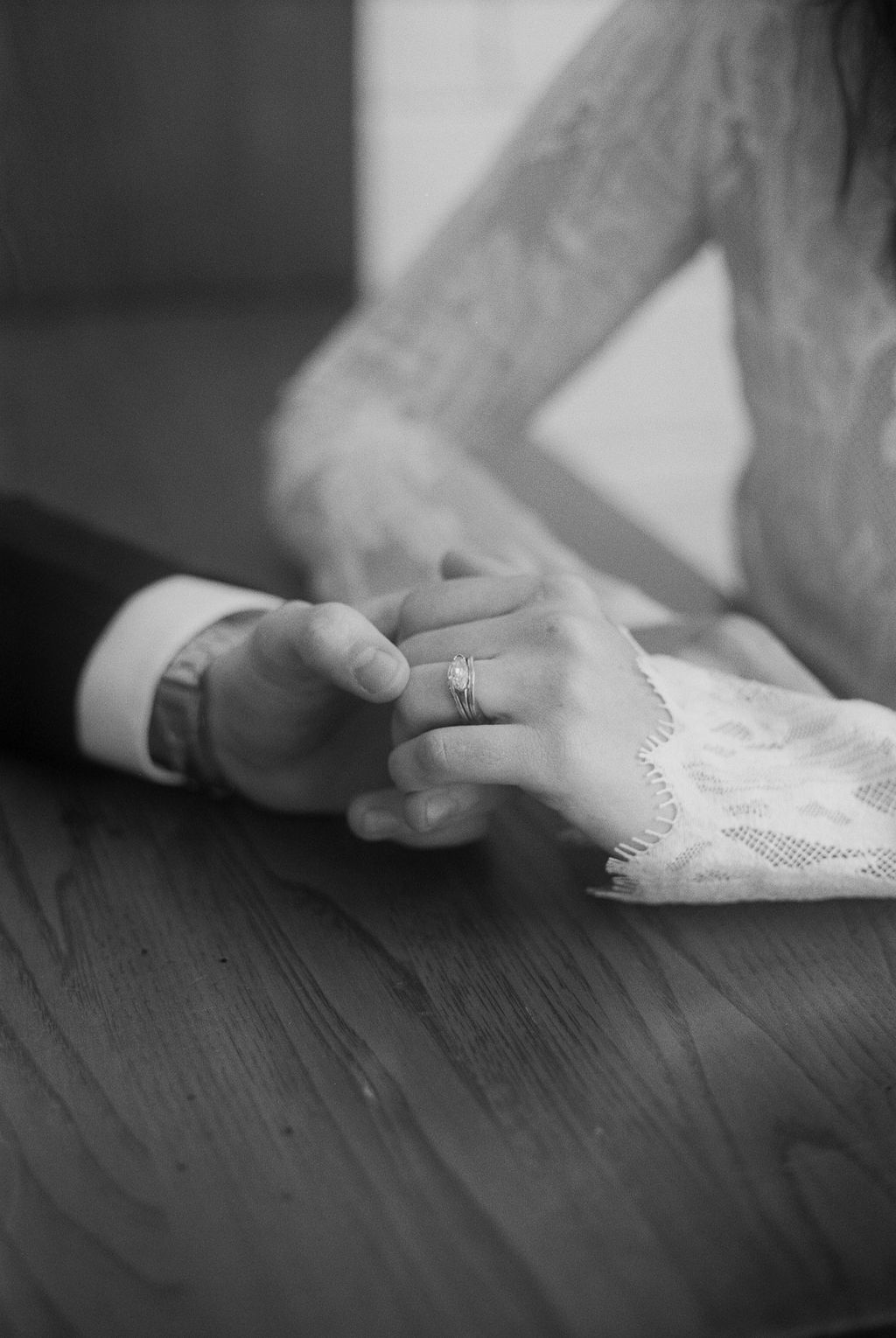 Close-up black-and-white photo of a couple holding hands on a wooden table, with a visible engagement ring on the woman's finger.
