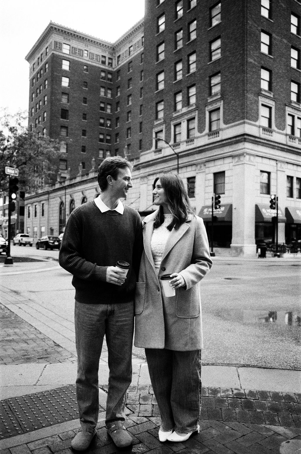 A smiling couple holding coffee cups, standing close together on a city sidewalk corner with tall buildings in the background.