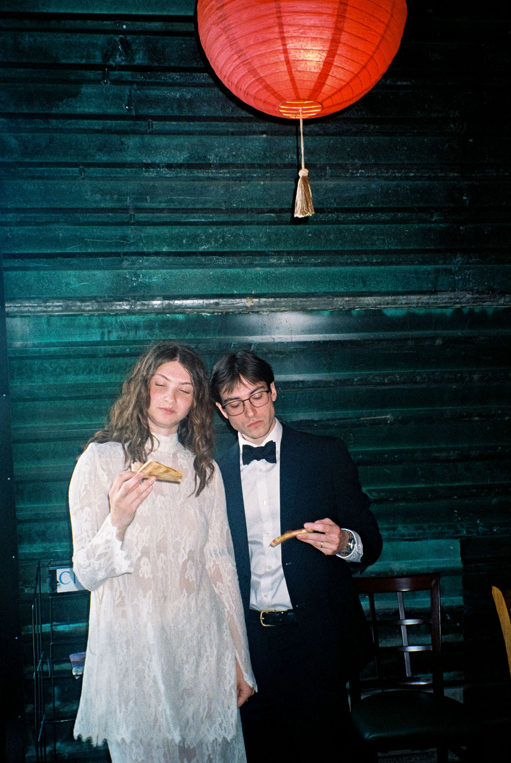 Man in a tuxedo and woman in a white lace dress standing indoors, each holding a slice of pizza, with a red paper lantern hanging above them.