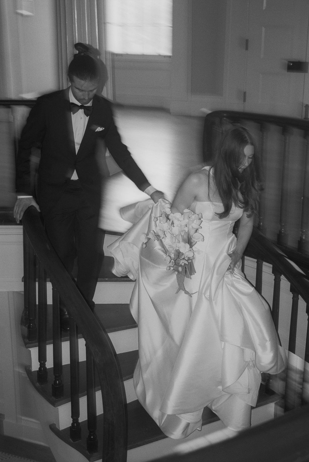 Black and white photo of a bride in a satin gown holding flowers and a groom in a tuxedo descending a staircase together.