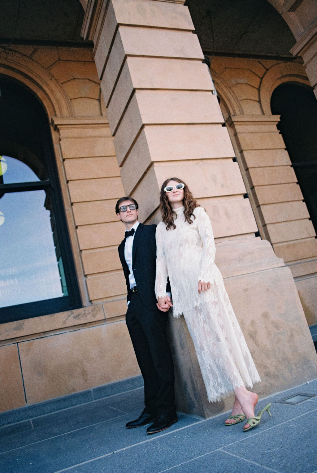 Man in a black suit and bow tie and woman in a white lace dress and green heels holding hands, leaning against a large stone pillar.