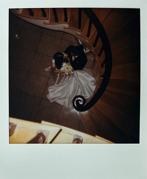 Top-down view of a bride in a white gown holding a bouquet and a groom in a black suit sitting together on a spiral wooden staircase.