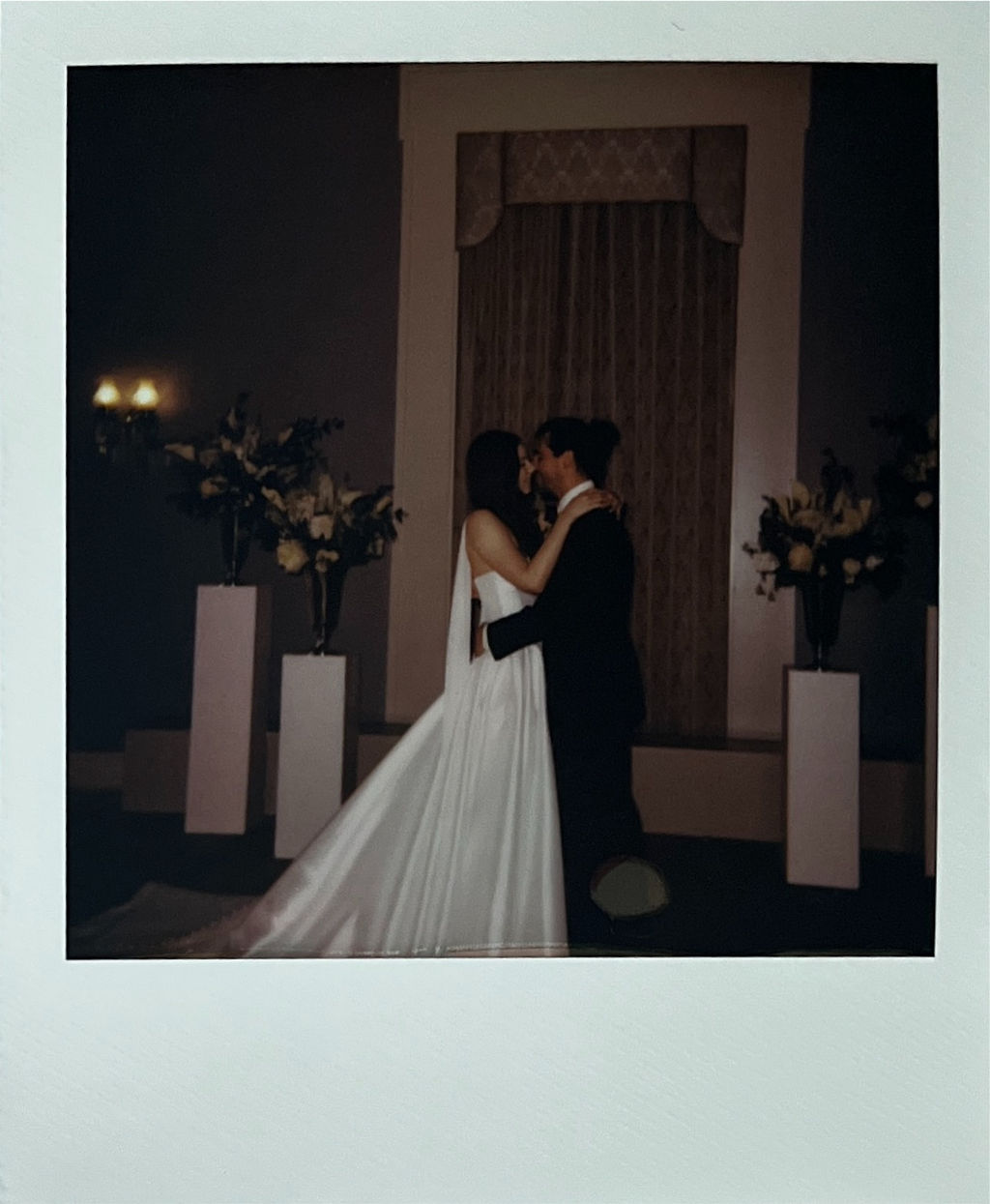 Bride and groom embracing and about to kiss in a dimly lit wedding venue with floral arrangements on pedestals.