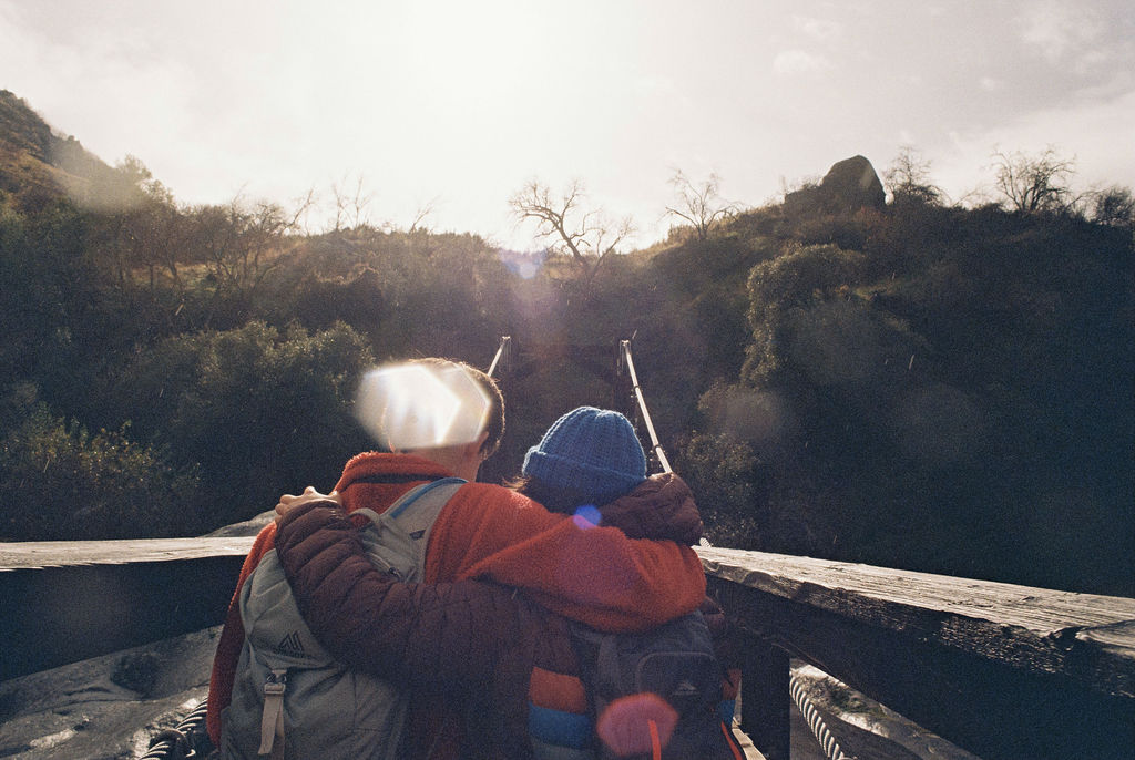 Two hikers with backpacks and arm around each other standing on a bridge overlooking a wooded valley with hills in the background.